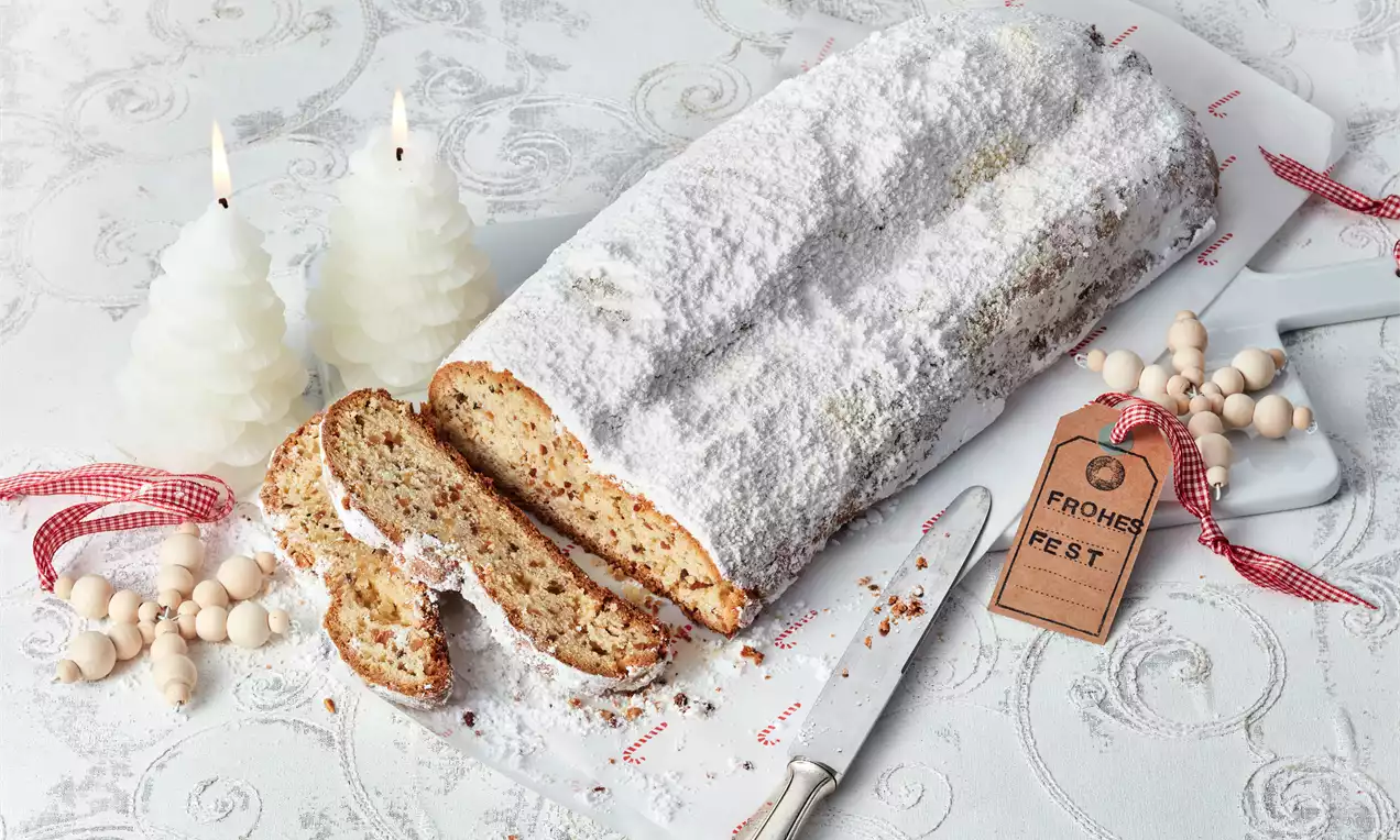 Goldbrauner Mandel-Stollen mit Puderzucker bestäubt, angeschnitten auf weißer Platte mit Holzperlen, rotem Band und Kerzen dekoriert