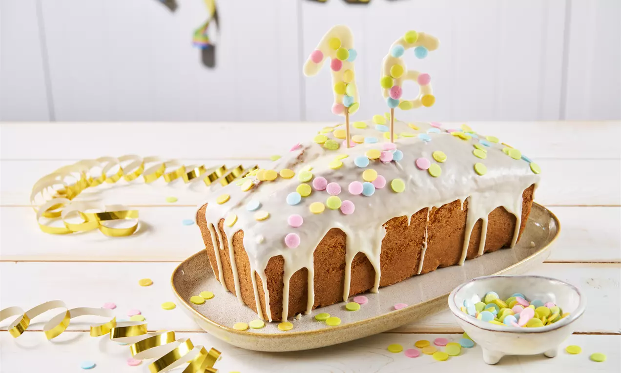Rechteckiger Rührkuchen mit weißer Glasur, bunten Konfettistreuseln und Zahlenkerzen, dekoriert auf einer goldenen Platte mit Partyaccessoires