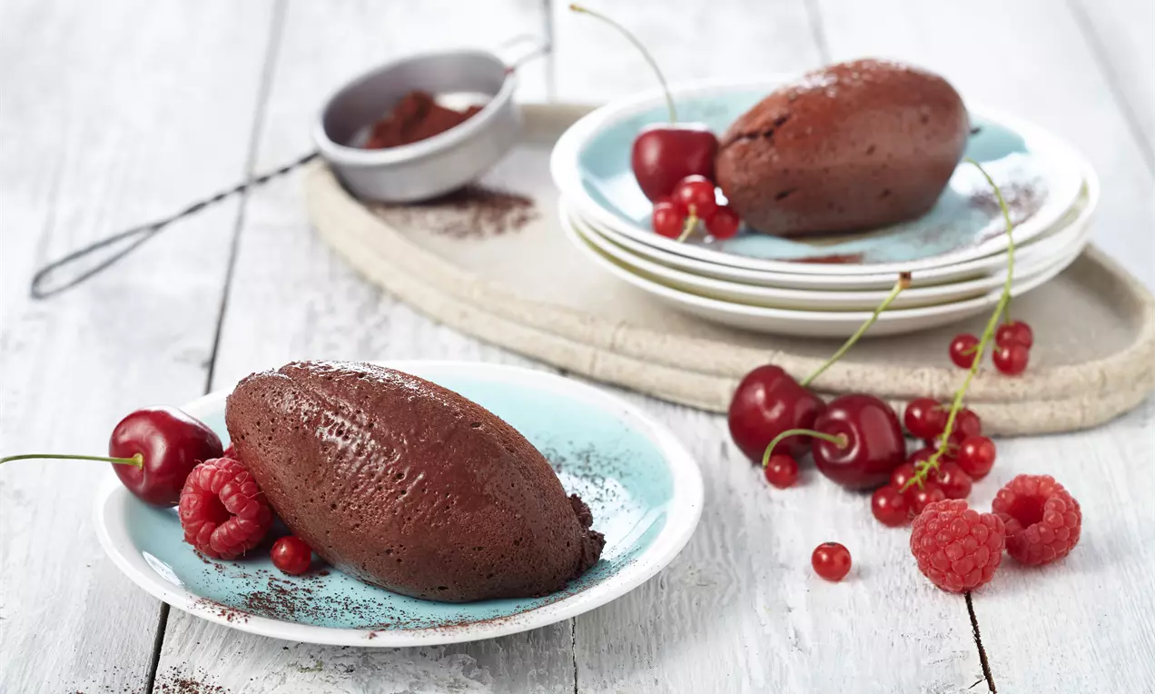 Schokoladen-Mousse in länglicher Form auf hellblauem Teller angerichtet, umgeben von Himbeeren, Kirschen und Kakao-Dekor