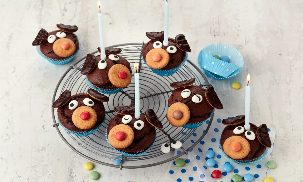 Schokoladenbraune Hundemuffins mit bunten Nasen, weißen Augen und Ohren aus Schokolade auf einem runden Gitter mit Kerzen verziert