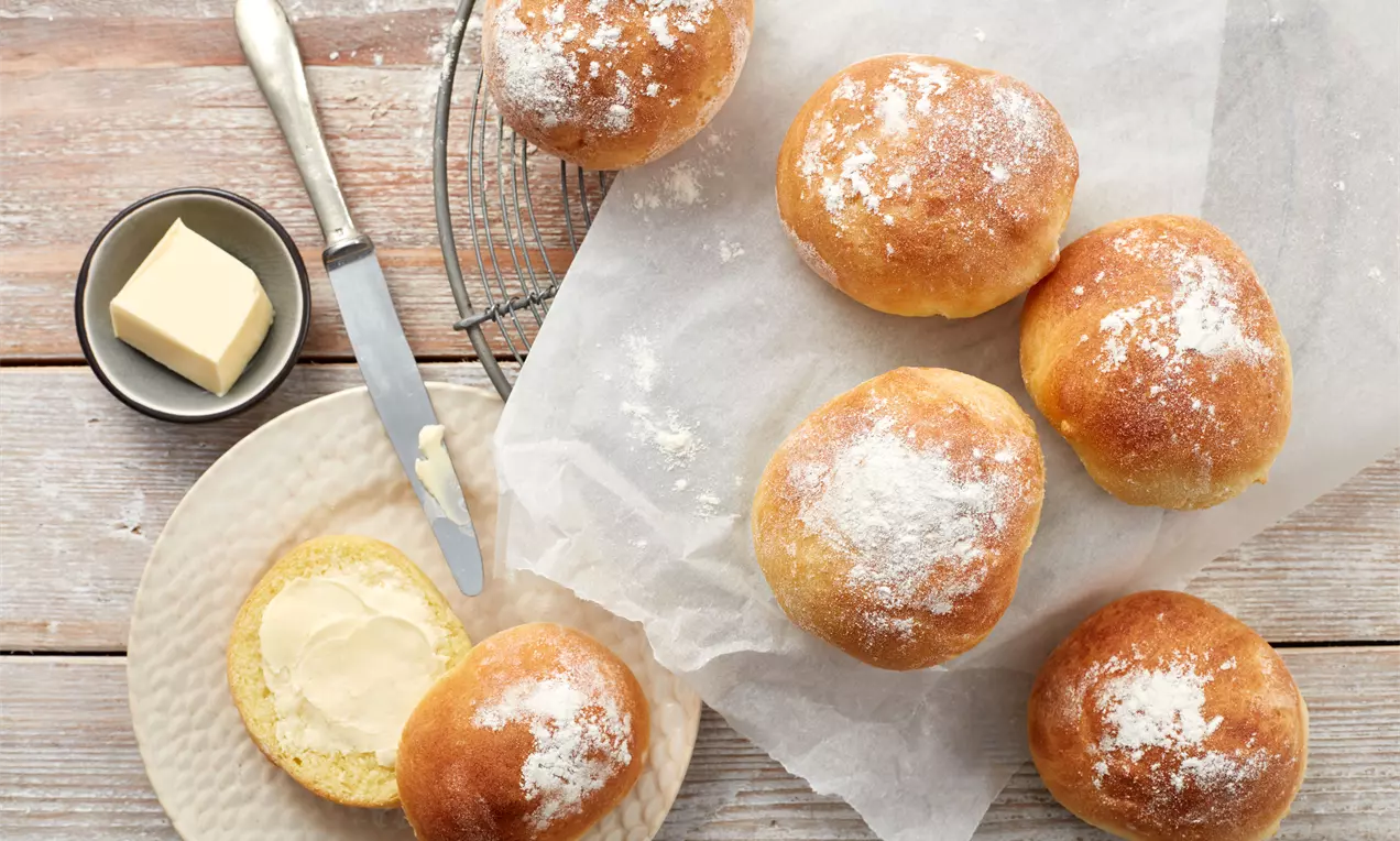 Goldbraune, runde Brötchen mit Mehlschicht auf Backpapier und Gitter, daneben ein aufgeschnittenes Brötchen mit Butter auf einem Teller