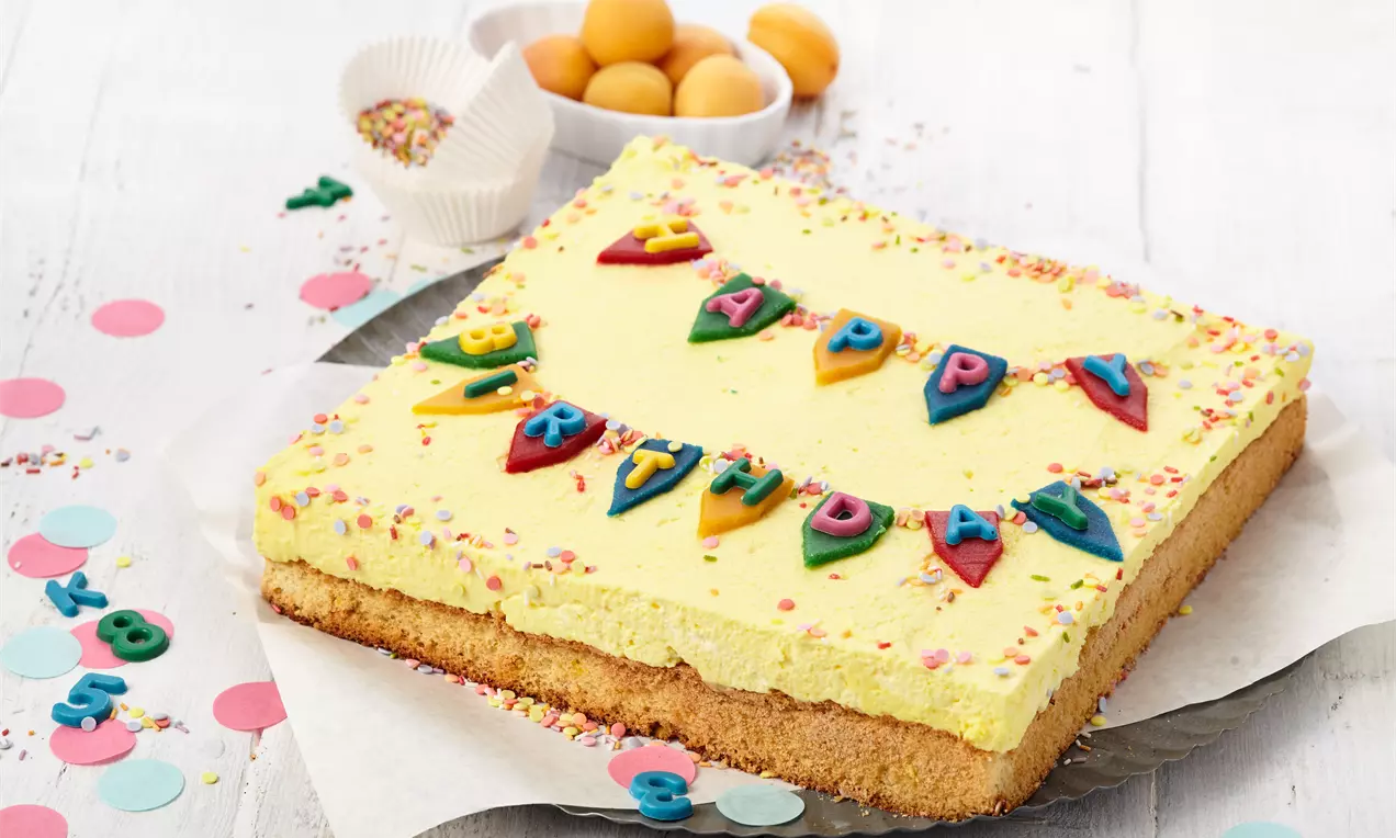 Quadratischer Geburtstagskuchen mit gelber Creme, bunter Zuckerdekoration, Happy-Birthday-Girlande und frischen Aprikosen im Hintergrund