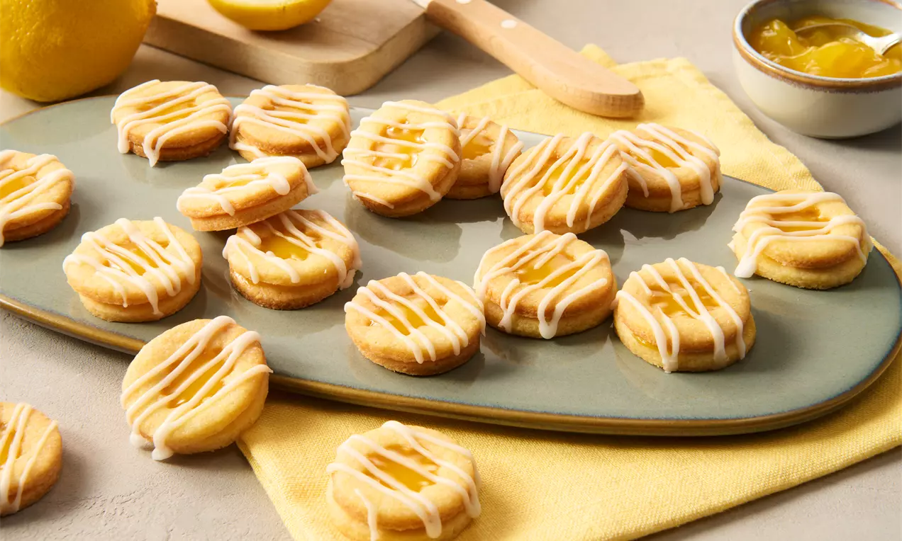 Runde Zitronenplätzchen mit gelber Füllung und weißem Zuckerguss auf länglicher Platte, dekoriert mit Zitronen und gelbem Tuch