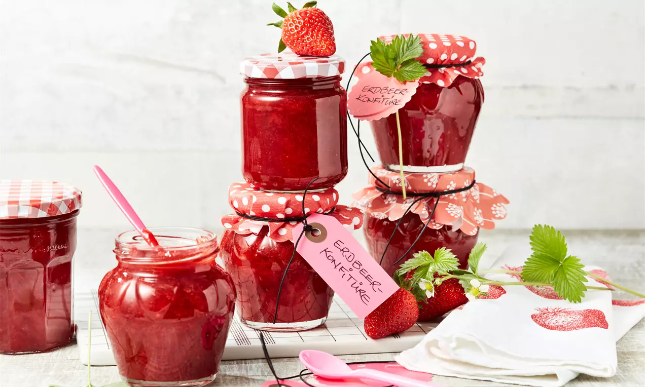 Erdbeer-Konfitüre in roten, glänzenden Gläsern mit karierten Deckeln, frischen Erdbeeren, grünem Blattwerk und rosa Löffeln arrangiert