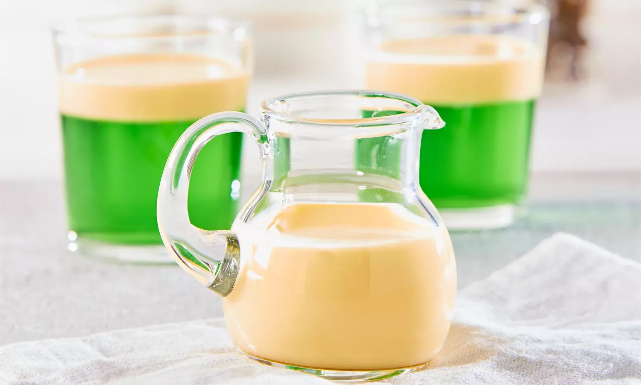 Kleine Glas-Karaffe mit cremig-beiger Vanillesoße auf einem hellgrauen Tuch, im Hintergrund grüne Desserts mit heller Schaumkrone