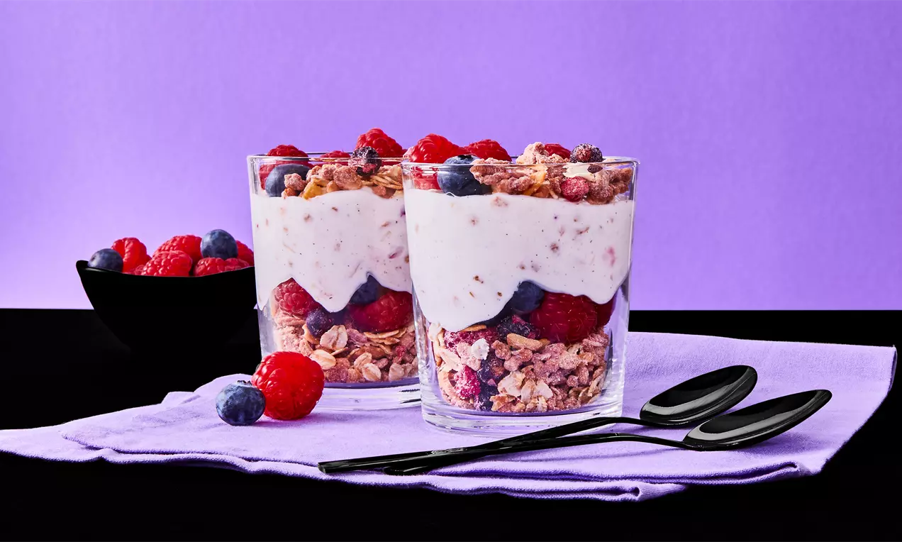 Zwei Gläser mit Schichten aus knusprigem Müsli, roten Beeren und cremigem Joghurt, arrangiert auf einem hellvioletten Tuch mit schwarzen Löffeln