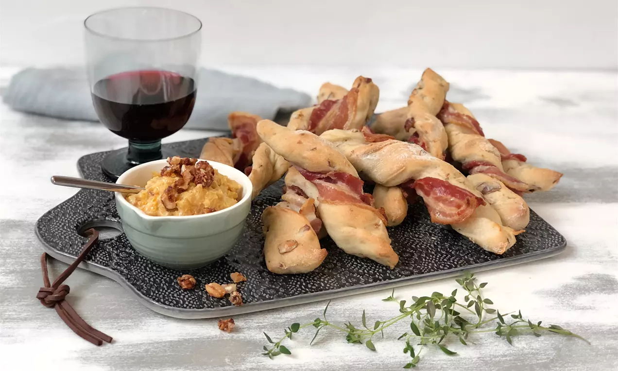 Gedrehte, goldbraune Brotsticks mit rotem Bacon umwickelt auf einer grauen Platte, serviert mit Dip in heller Schale und frischem Thymian