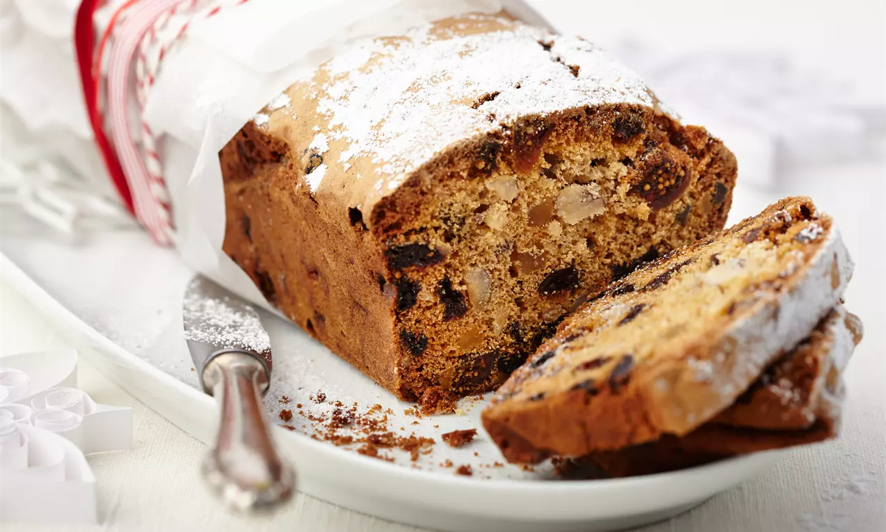 Früchtekuchen mit goldbrauner Kruste, eingebackenen Früchten und Mandeln, leicht mit Puderzucker bestäubt auf weißem Teller mit Messer