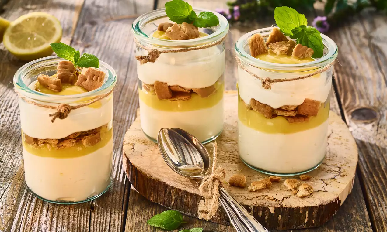 Quark-Schichtdessert im Glas mit cremig-weißer Quarkmasse, gelbem Lemon Curd, Keksstücken und frischer Minze auf Holzscheibe angerichtet
