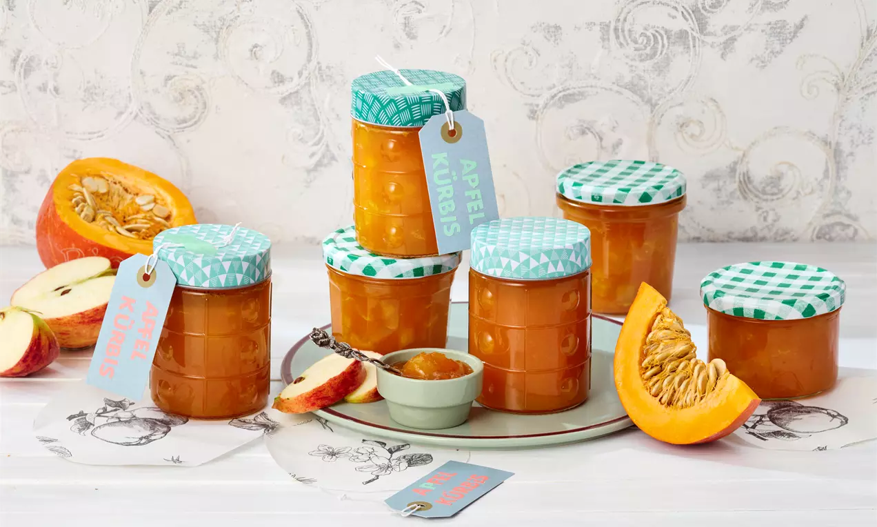 Gläser mit goldorangener Marmelade und grün gemusterten Deckeln, umgeben von Apfel- und Kürbisstücken auf hellem Untergrund