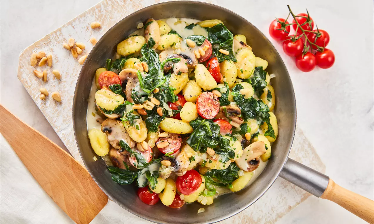 Gelblich-goldene Gnocchi mit grünem Spinat, roten Kirschtomaten und braunen Champignons in einer silbernen Pfanne angerichtet