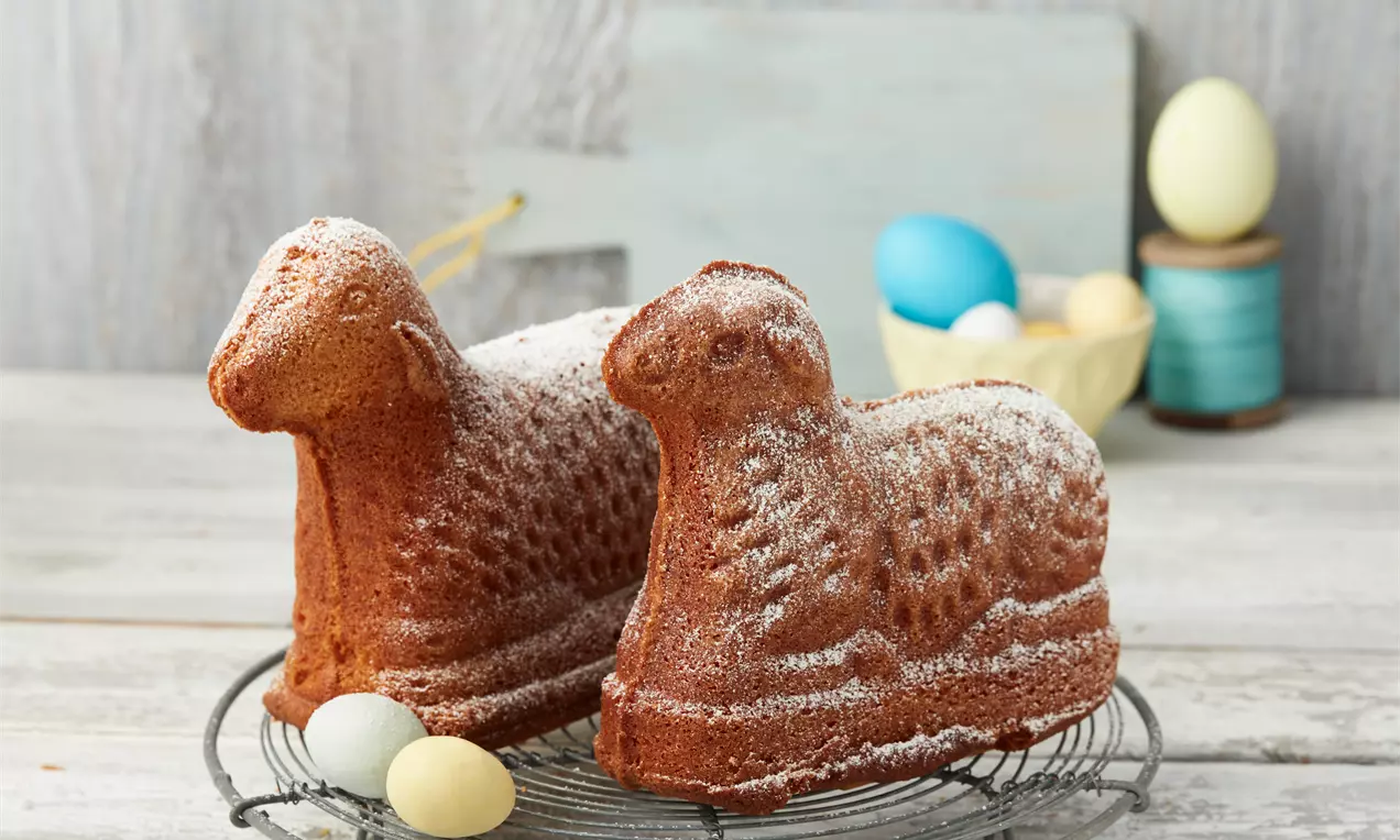 Zwei goldbraune Osterlämmer aus Rührteig mit Puderzucker bestäubt auf einem runden Metallgitter, umgeben von bunten Eiern und Dekorationen