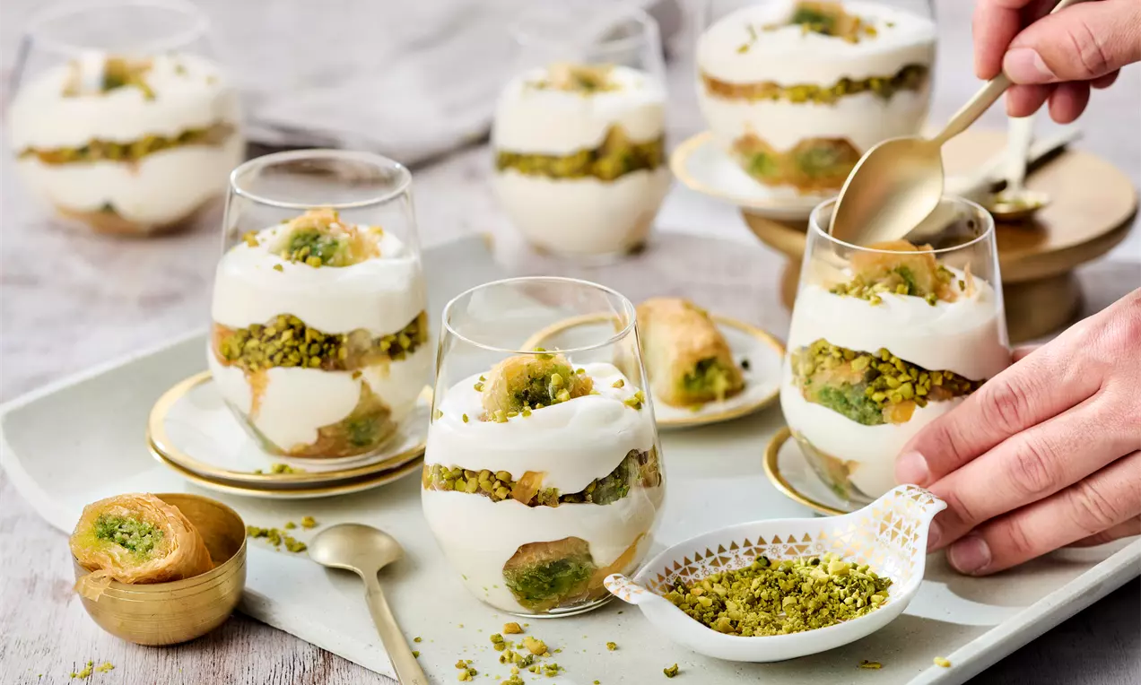 Schichtdessert in Gläsern mit weißer Creme, goldbraunem Gebäck und grünen gehackten Pistazien, angerichtet auf hellem Tablett mit Löffeln