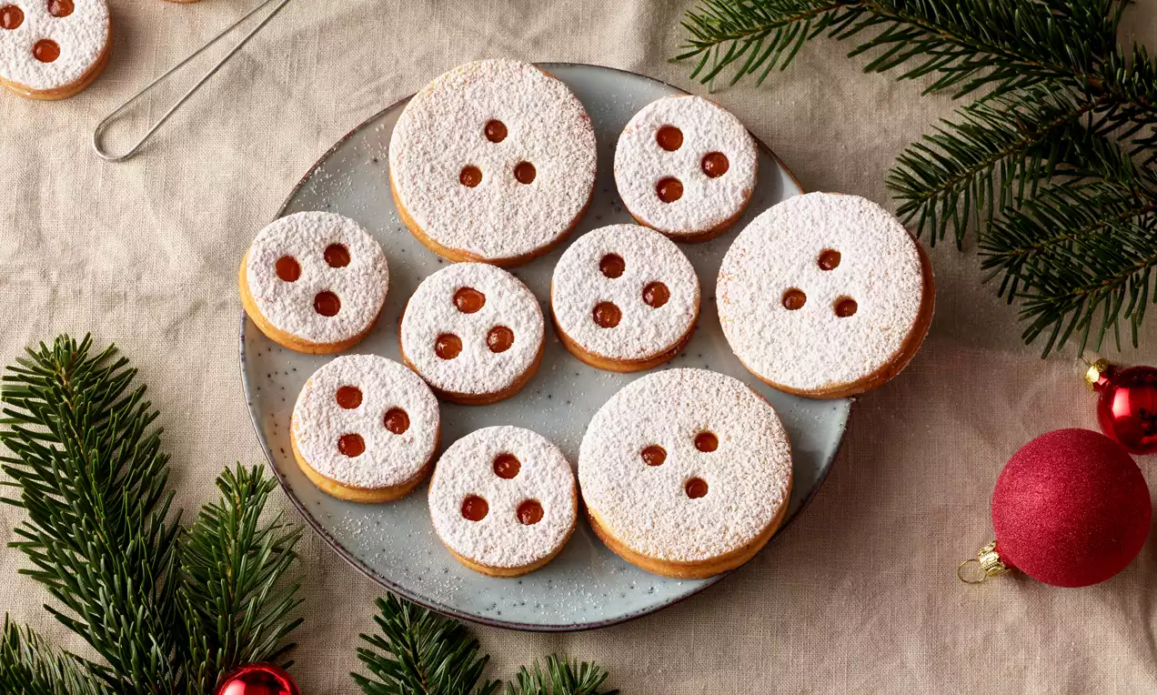 Linzer Augen mit goldbraunen Keksböden und weißem Puderzucker bestäubt, gefüllt mit roter Marmelade, auf grauem Teller mit Tannenzweigen und Weihnachtskugeln