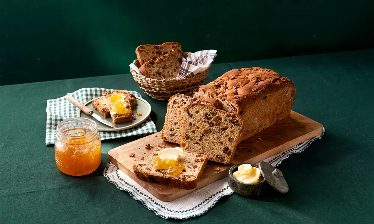 Irisches Früchtebrot mit goldbrauner Kruste, samtigen Scheiben voller Früchte, arrangiert auf Holzbrett mit Butter, Marmelade und grünem Tuch