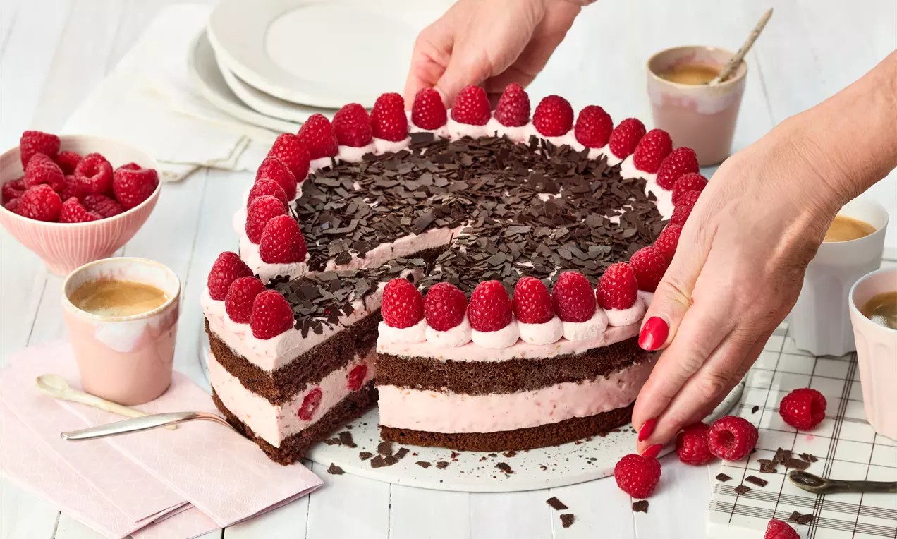 Schoko-Himbeer-Torte mit dunklen Böden, heller Himbeercreme, frischen Himbeeren und Schokoraspeln, dekorativ angerichtet auf weißem Teller