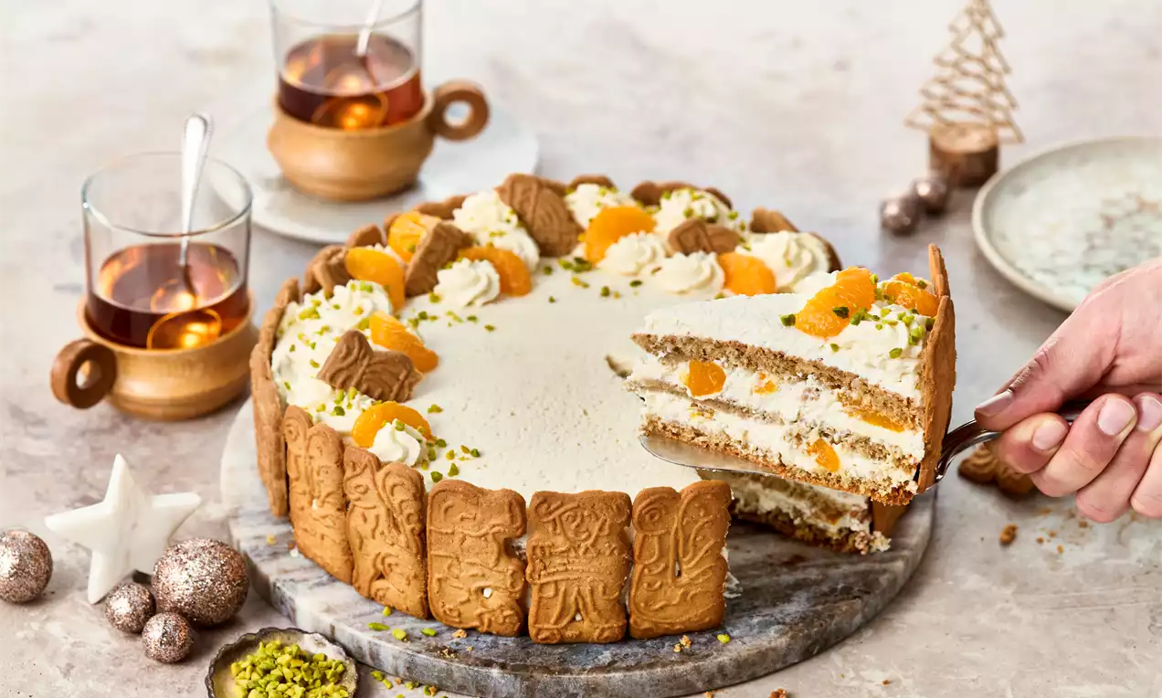 Spekulatius-Torte mit Keksrand, luftiger Sahnefüllung, Mandarinenstückchen, Pistazien und Keksen auf Marmorteller, daneben Tee in Holzbechern