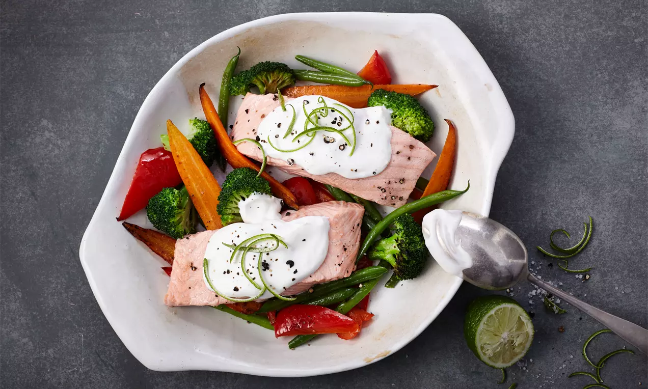 Zartrosa Lachs mit weißer Creme und grünen Limettenzesten auf gebratenem Gemüse in kräftigen Farben in einer weißen Schale angerichtet