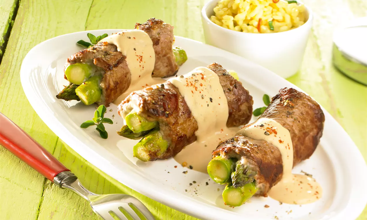 Gebratene Roastbeef-Röllchen mit grünen Spargelspitzen gefüllt, hellbrauner Sauce überzogen, auf weißem Teller mit Kräuter-Dekoration angerichtet