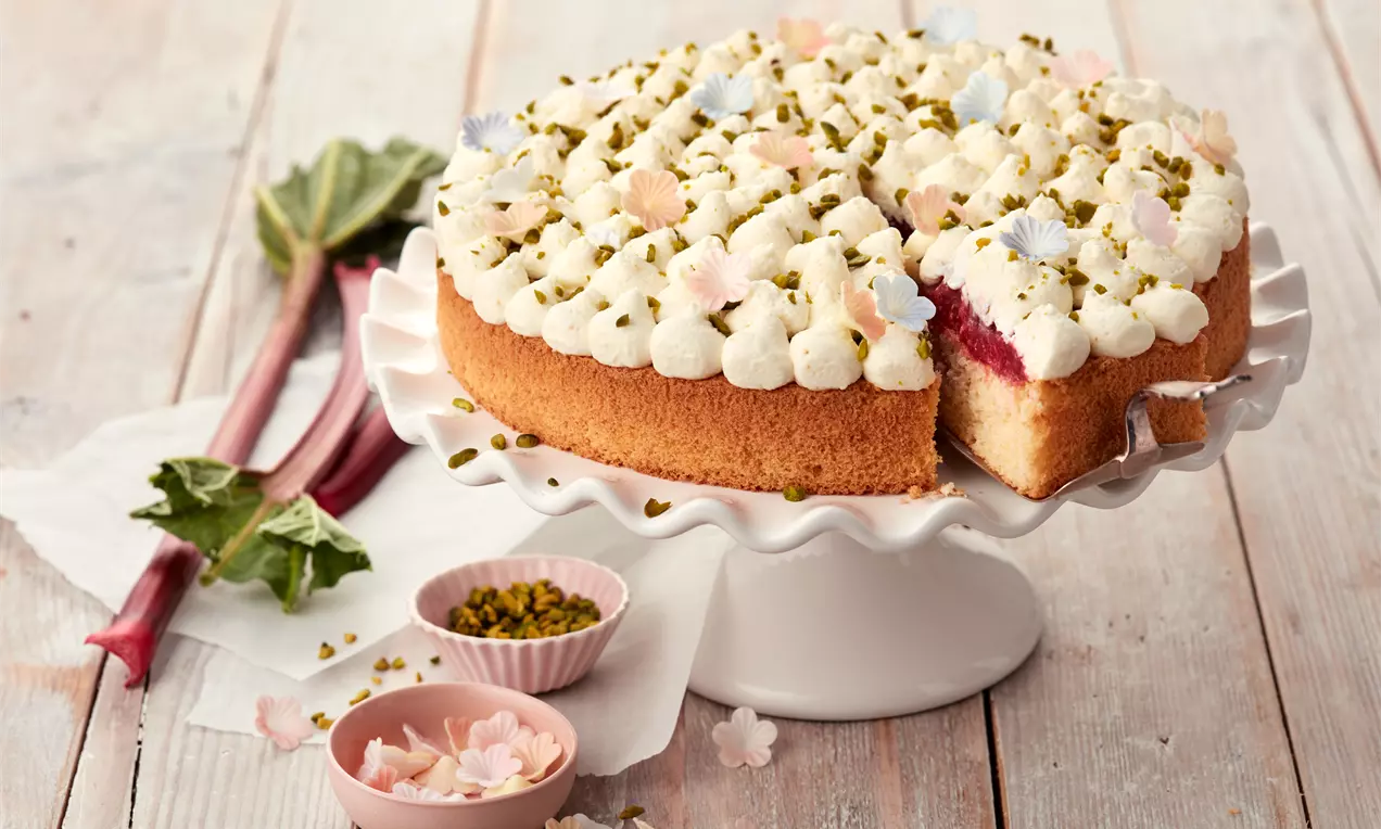 Runde Rhabarber-Torte mit goldenem Biskuitboden, weißer Sahnetupfen-Dekoration, gehackten Pistazien und Blüten auf einer weißen Tortenplatte