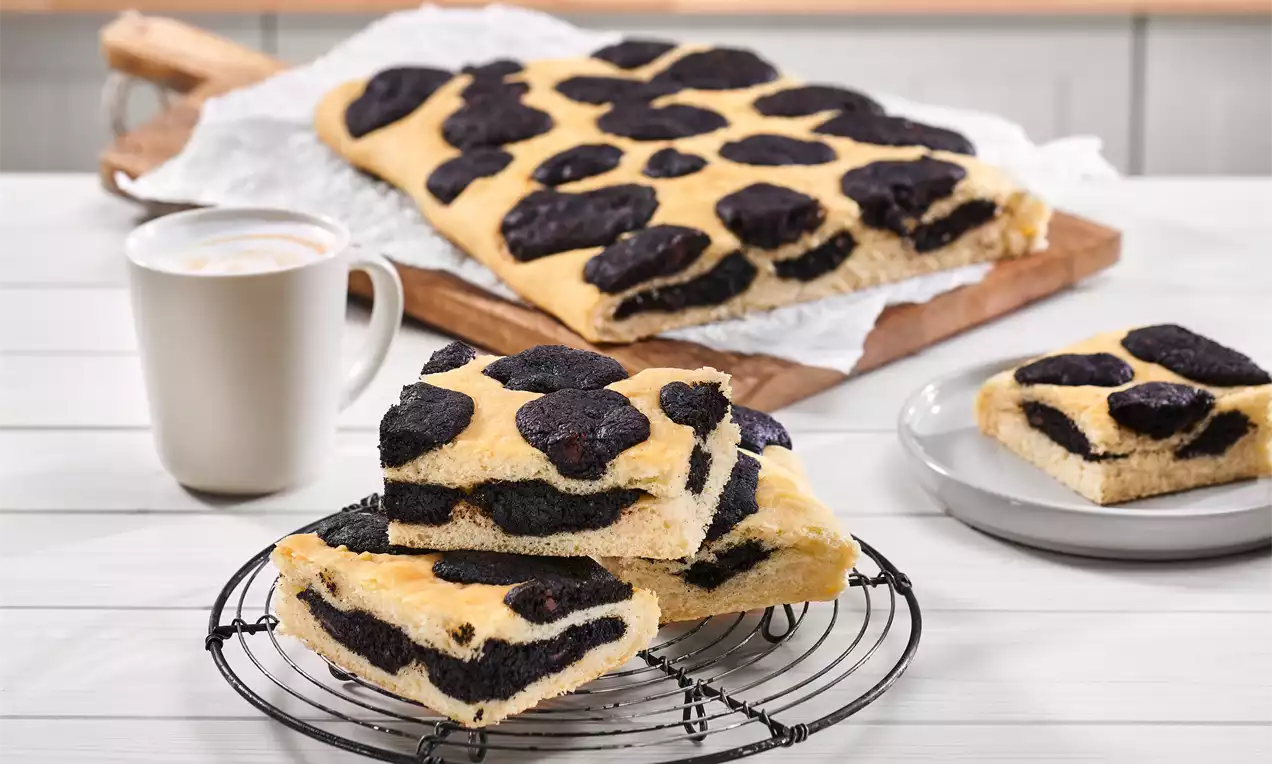 Goldbraune Cookie-Focaccia mit schwarzem Keksmuster auf Gitter, daneben ein Stück auf weißem Teller und eine Tasse Cappuccino.