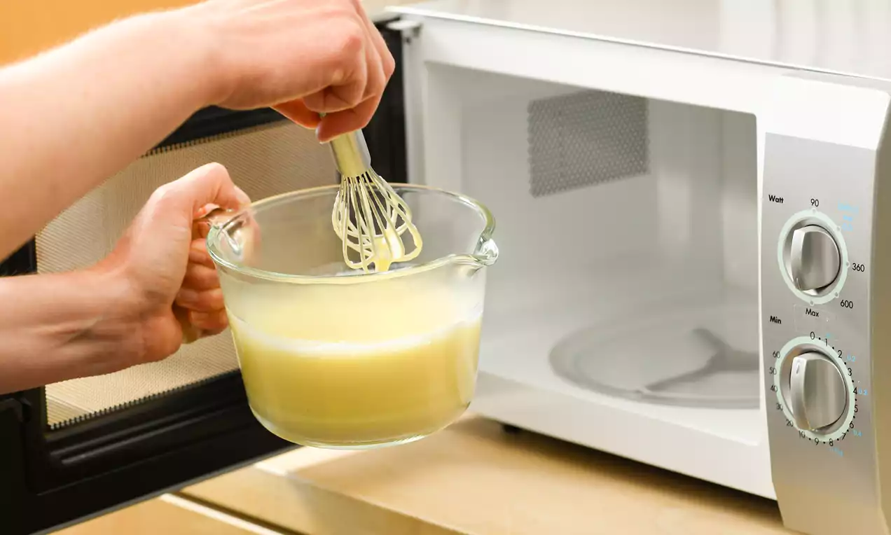 Eine transparente Glasschüssel mit gelblich-cremigem Pudding wird mit einem Schneebesen vor einer geöffneten Mikrowellentür verrührt