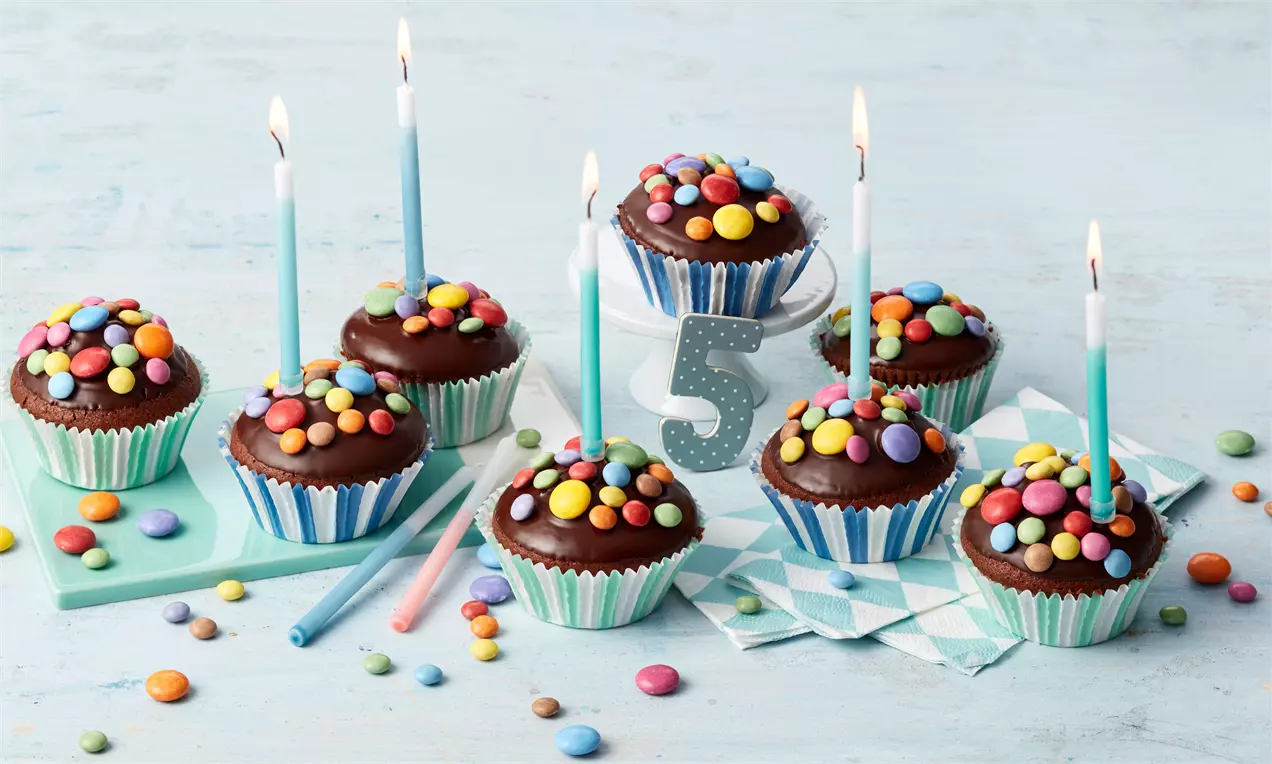 Schokoladenmuffins in gestreiften Förmchen mit bunter Smarties-Deko, Kerzen und einer funkelnden fünf auf hellblauer Tischkante