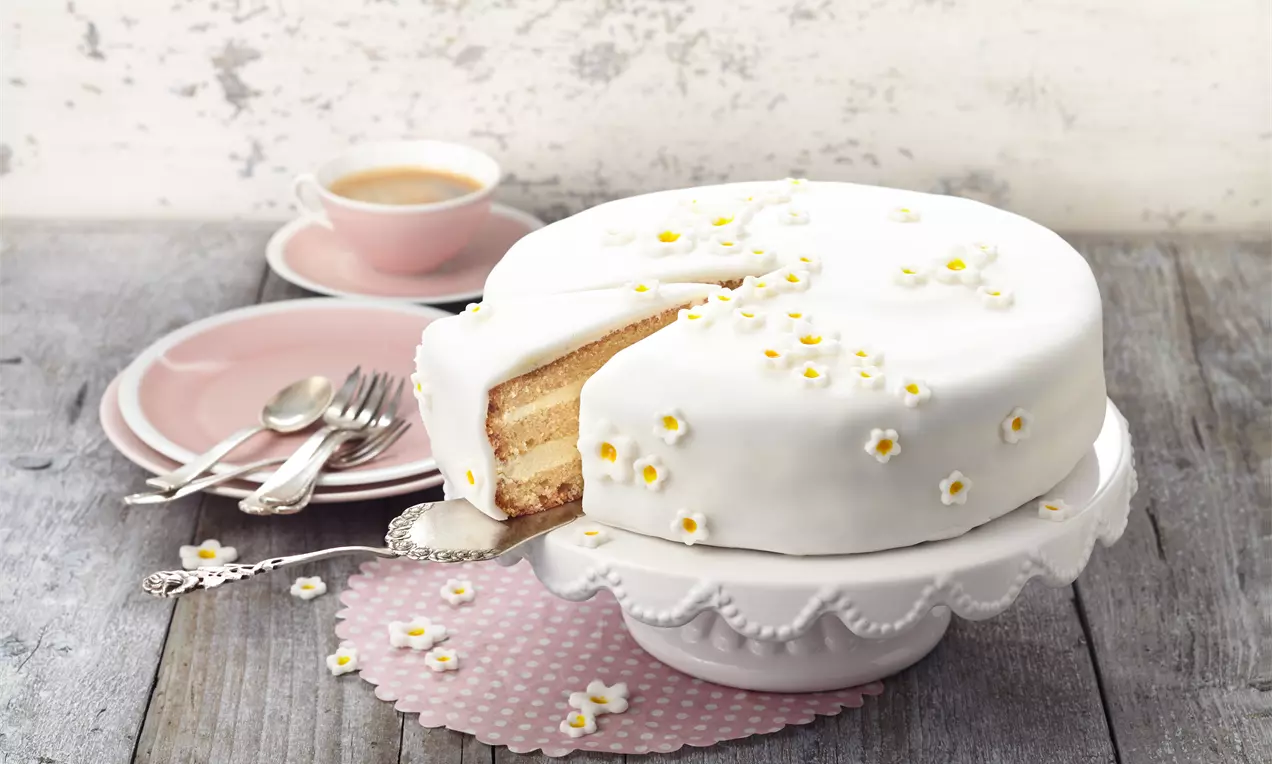 Runde weiße Torte mit glatter Fondant-Decke und kleinen gelben Zuckerblüten, angeschnitten auf einer weißen Tortenplatte mit rosa Geschirr im Hintergrund