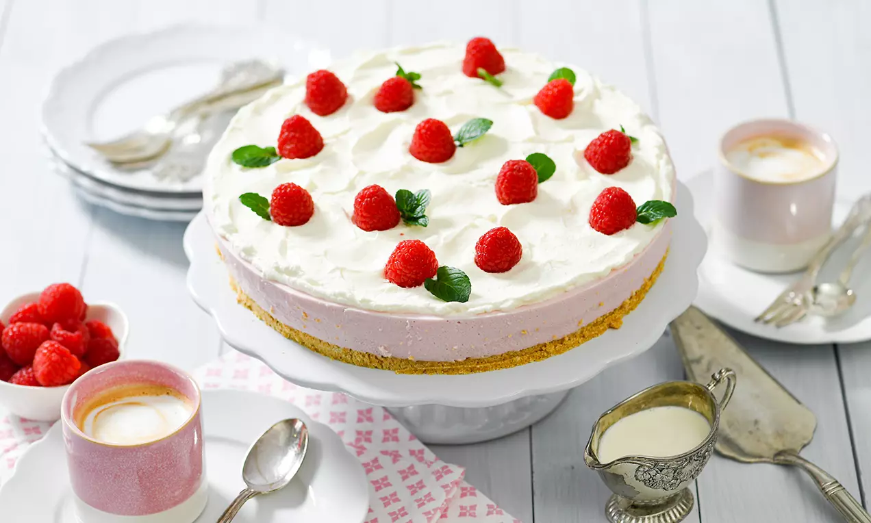 Frischkäse-Torte mit rosa Himbeerfüllung auf Keksboden, getoppt mit weißer Sahnehaube, frischen Himbeeren und Minzblättern auf einer Tortenplatte