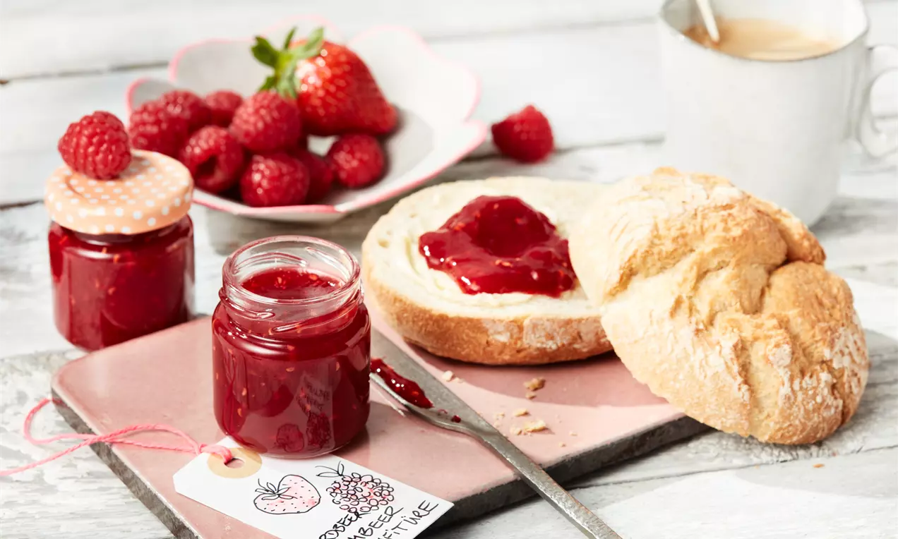 Erdbeer-Himbeer-Konfitüre mit tiefroter, glänzender Textur in offenem Glas neben einem Brötchen mit Konfitüre auf hellem Tablett