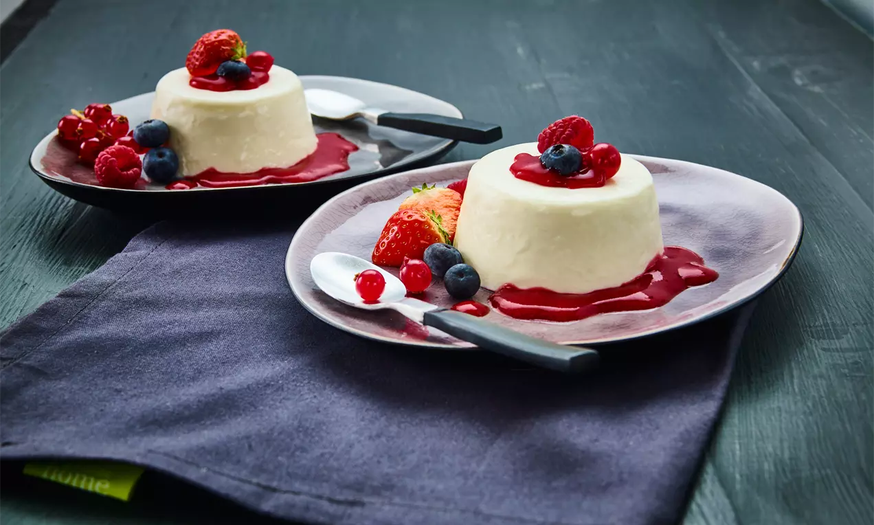 Weiße gestürzte Quarkcreme mit glatter Oberfläche, serviert auf einem flachen Teller, garniert mit roten Beeren und Beeren-Sauce