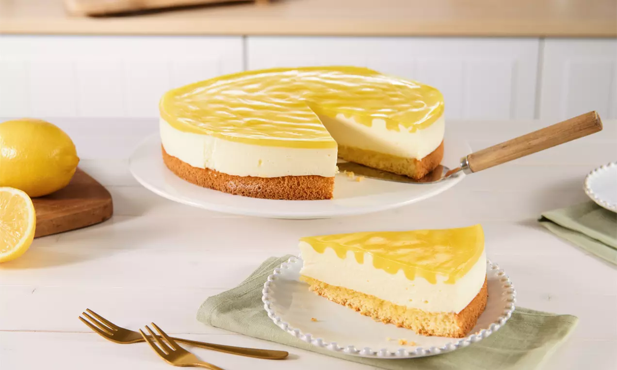 Zitronen-Joghurt-Torte mit glänzend gelber Creme auf lockerem Keksboden, dekorativ auf einer weißen Platte mit angeschnittenem Stück
