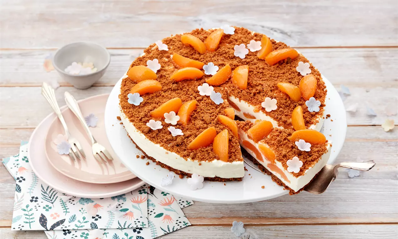 Käsekuchen mit cremiger weißer Füllung, braunem Keksboden, Aprikostückchen und dekorativen Blüten aus Zucker auf weißer Tortenplatte