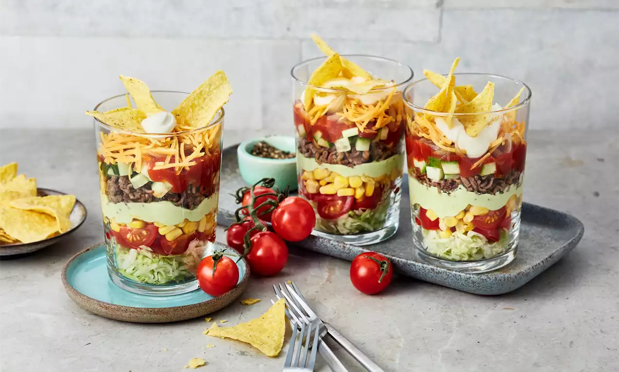 Schichtsalat in klaren Gläsern mit frischen Zutaten wie Mais, Tomaten, Avocadocreme, geriebenem Käse und Tortillachips, dekoriert mit Kirschtomaten