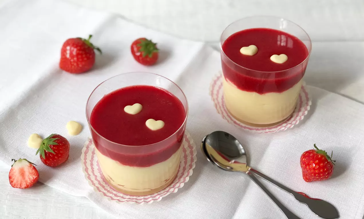Feiner, hellgelber Pudding in klaren Gläsern, bedeckt mit roter Erdbeersoße und weißen Schokodekoren, arrangiert auf weißem Baumwolltuch