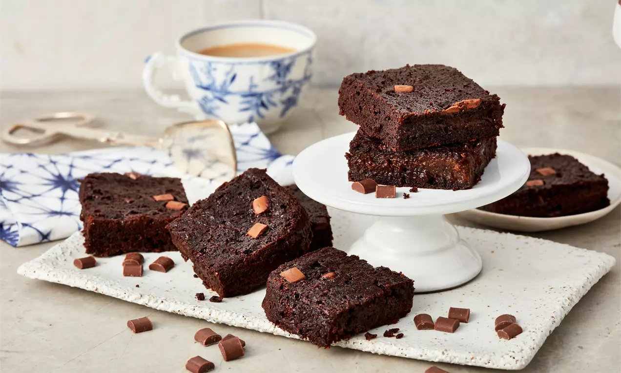 Dunkle, saftige Süßkartoffel-Brownies mit Schokostückchen, teils gestapelt auf weißer Etagere, daneben verstreute Schokowürfel und eine Tasse Tee