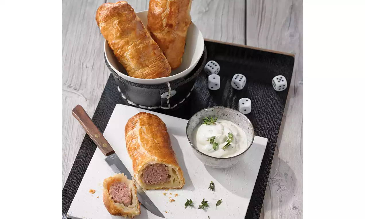 Goldbraune Mettrollen mit knuspriger Blätterteighülle, eine aufgeschnitten mit sichtbarer rosa Füllung, angerichtet mit Kräuterdip und Messer