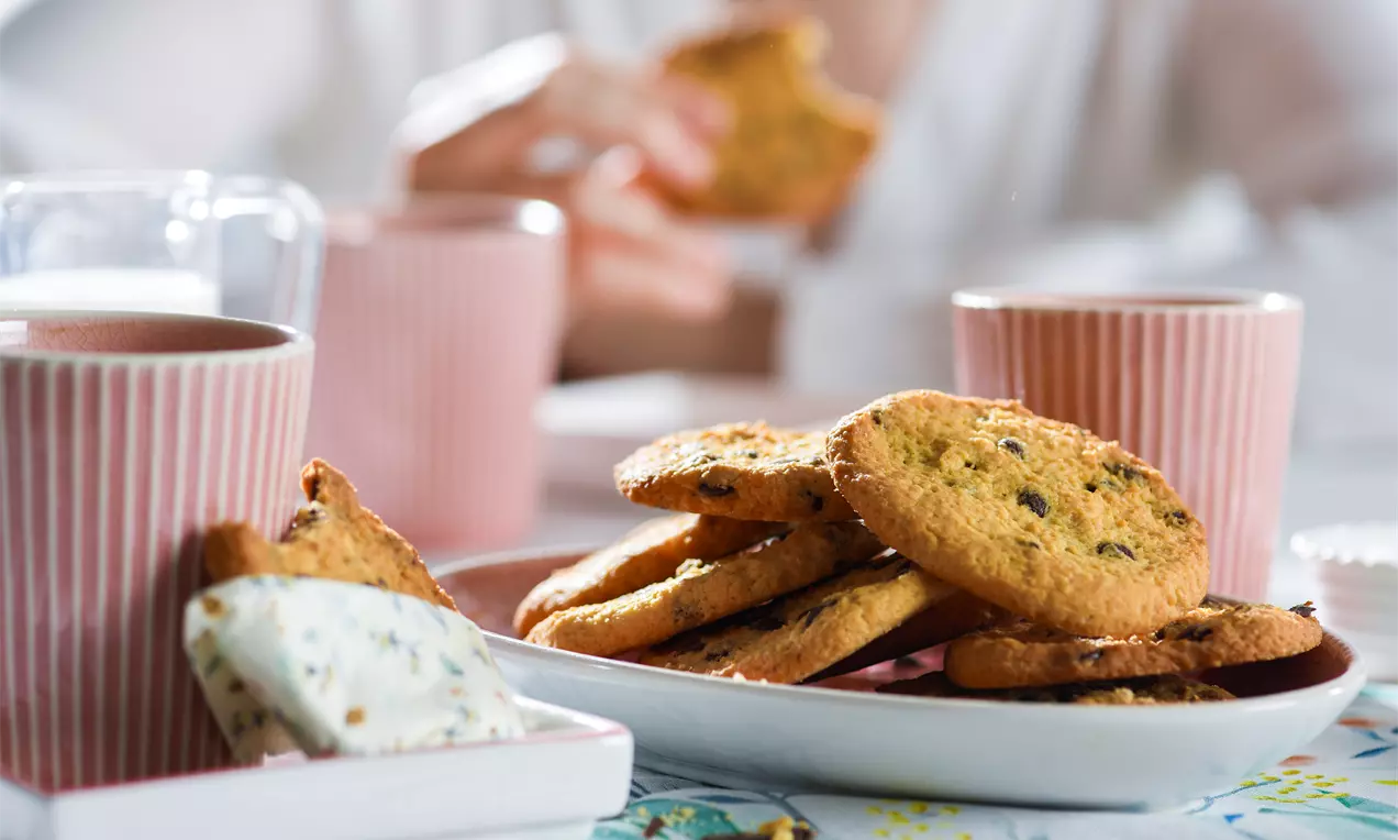 Goldene Low-Carb-Plätzchen mit knuspriger Textur und Schokostückchen auf weißem Teller, daneben rosa Tassen und helle Dekoration