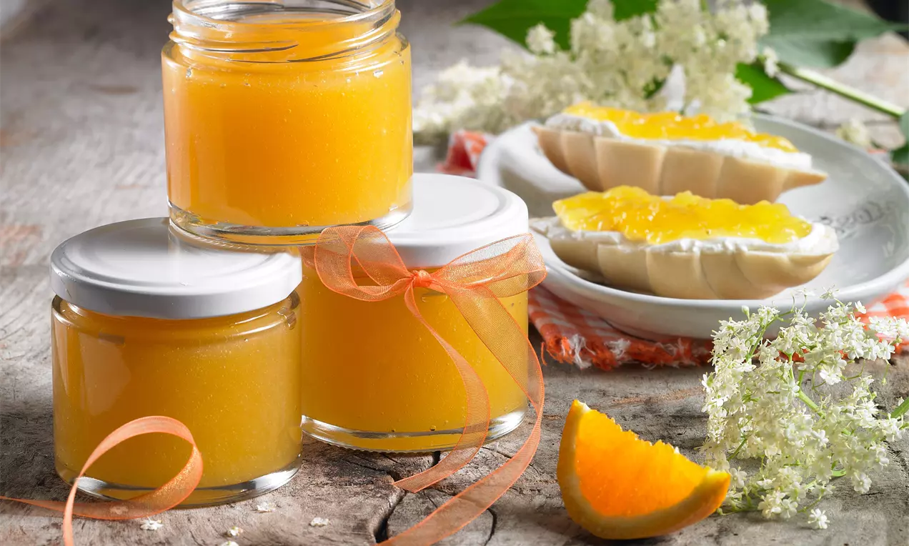 Drei Gläser mit leuchtend orangefarbenem Gelee, dekoriert mit Schleifen, daneben frische Holunderblüten und Marmeladenbrötchen auf rustikalem Holzbrett