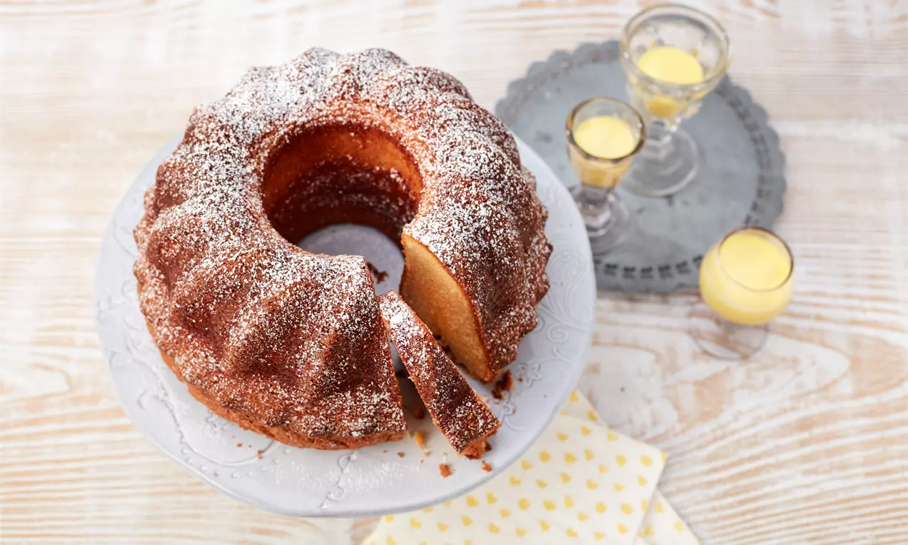 Saftiger Gugelhupf mit goldbrauner Kruste, leicht mit Puderzucker bestäubt, auf einer weißen Kuchenplatte neben Gläsern mit Eierlikör