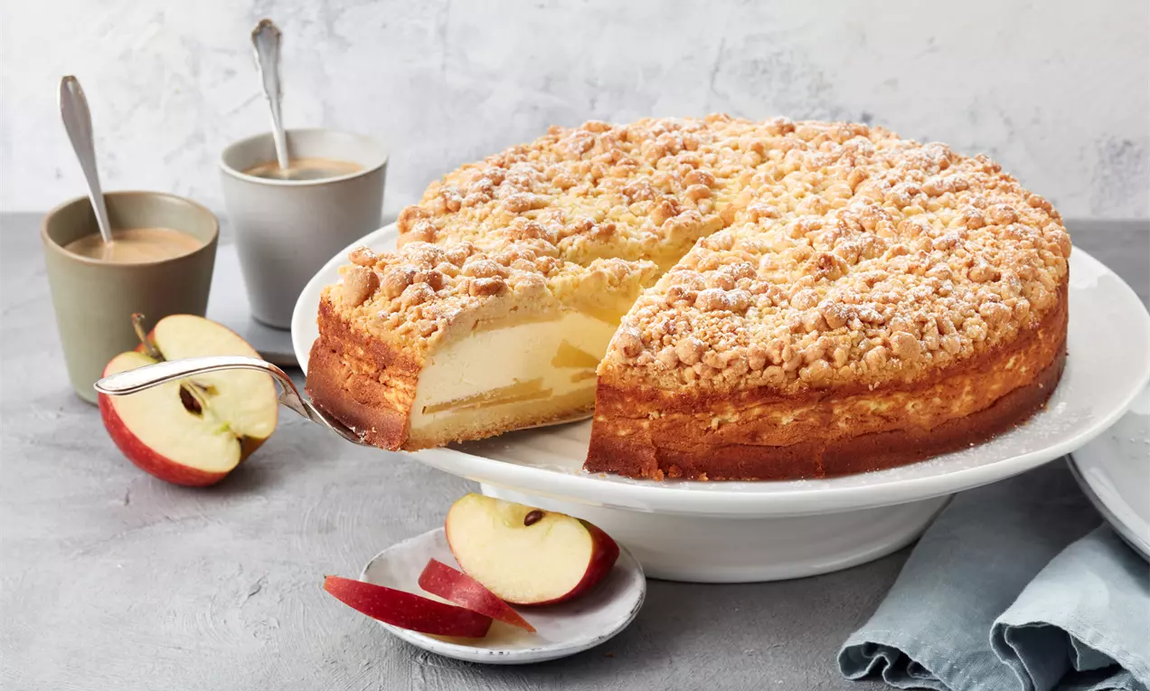 Apfel-Käsekuchen mit goldbrauner Streuselkruste und cremiger Füllung auf weißer Platte, dekoriert mit Apfelscheiben und zwei Tassen Kaffee