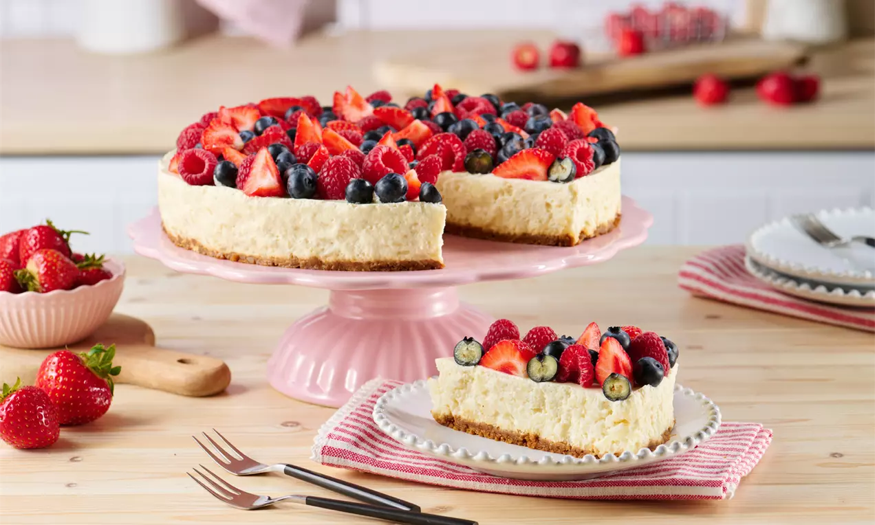 Milchreistorte mit goldbraunem Boden, cremiger Füllung, dicht belegt mit Erdbeeren, Himbeeren und Blaubeeren auf rosa Tortenplatte drapiert