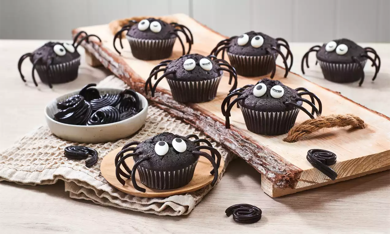Dunkle Schokoladenmuffins mit weißen Augen aus Zuckerglasur und schwarzen Lakritzbeinen auf einer Holzplatte arrangiert
