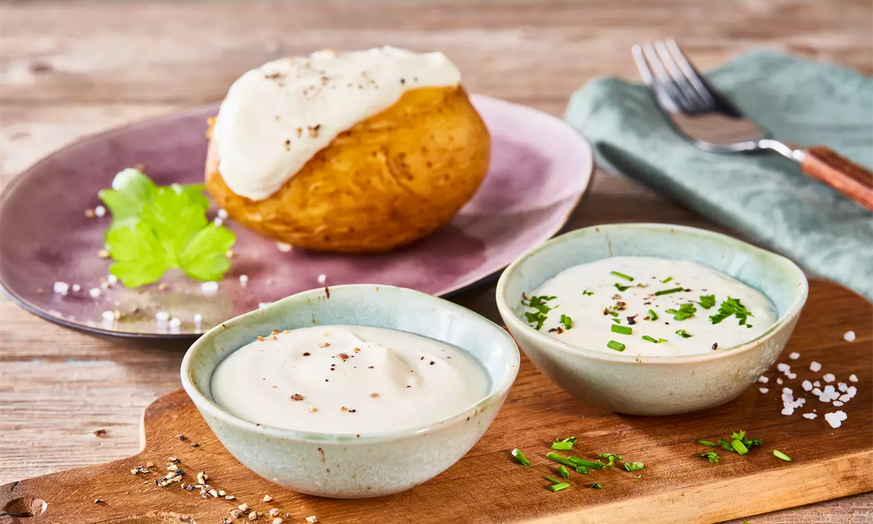 Goldene Ofenkartoffel mit weißer Sour Cream bestrichen auf einem violetten Teller, dazu zwei Keramikschälchen mit Kräuterdips auf Holzbrett