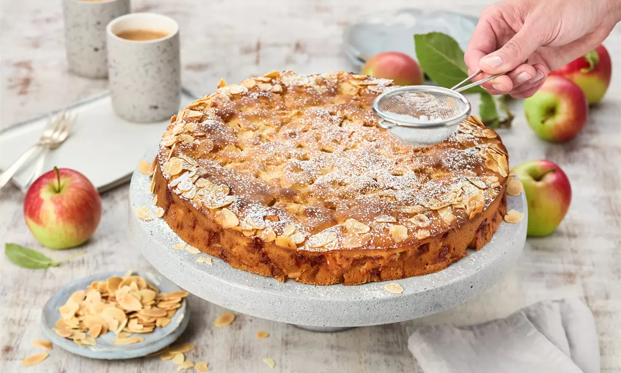 Goldbrauner Apfel-Joghurt-Kuchen mit Mandelblättchen und Puderzucker bestäubt auf einer hellen Kuchenplatte, umgeben von frischen Äpfeln