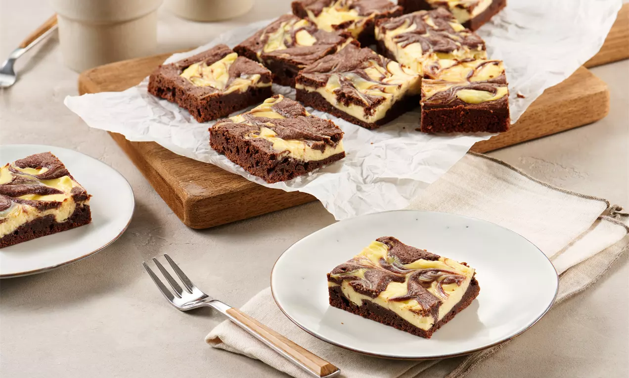 Cheesecake-Brownies mit schokoladigem Teig und heller Käsekuchen-Marmorierung auf Backpapier und Tellern angerichtet