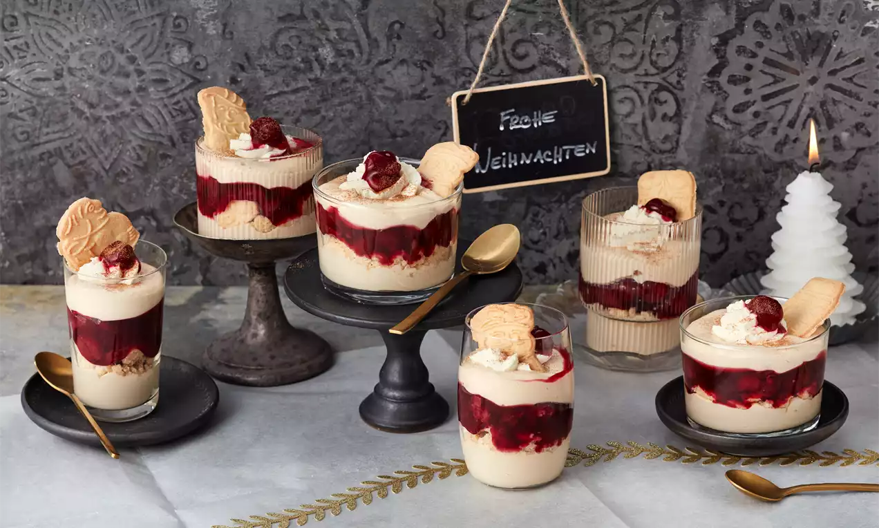 Dessertgläser mit cremefarbener Spekulatius-Mousse, dunkler Kirschschicht, Sahne und Spekulatius-Deko auf rustikalem Tisch drapiert