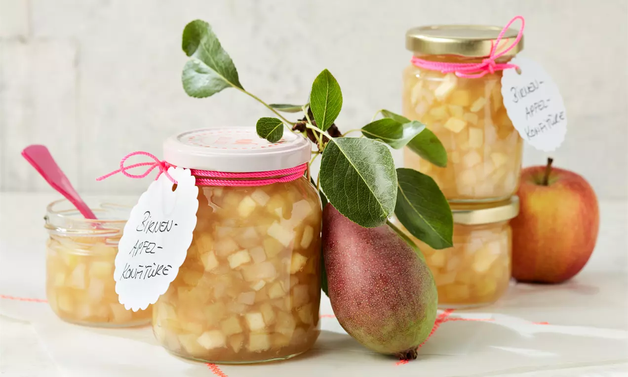 Glasgefäße mit goldgelber Birnen-Apfel-Konfitüre, umgeben von einer Birne, einem Apfel, grünen Blättern und einem pinken Löffel