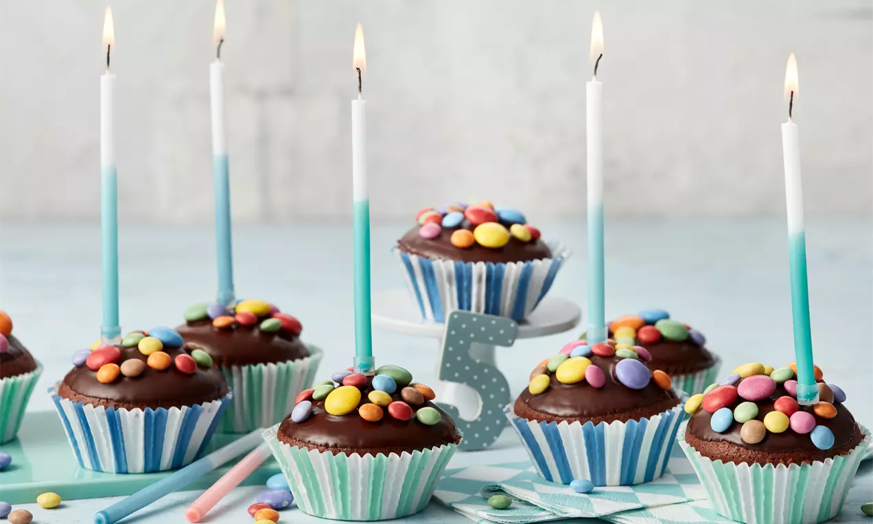 Schokoladenmuffins in gestreiften Papierförmchen, verziert mit bunten Schokolinsen und brennenden weißen Kerzen auf hellblauem Untergrund