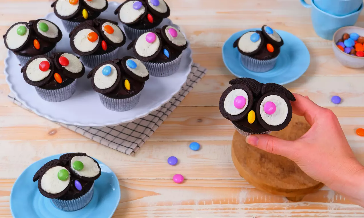 Eulen-Muffins mit schwarzem Teig, Oreo-Augen mit weißen Keksfüllungen und kleinen orangen Zuckerschnäbeln, auf einem Teller arrangiert